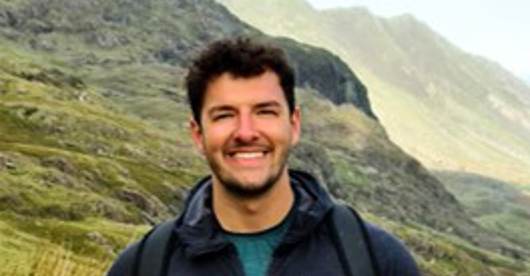 white man with blue jacket stood in front of mountain backdrop
