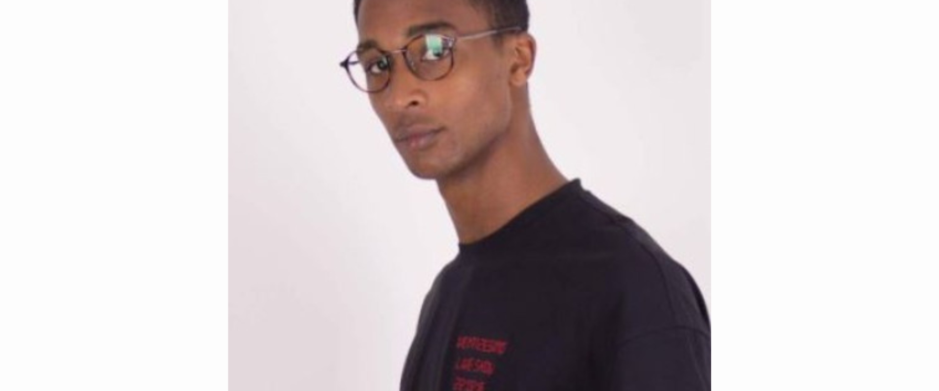 A person wearing a black T‑shirt with small red text on the front, shown from the shoulders up against a plain light background. The individual is standing at a slight angle, with the head turned partially to the side.