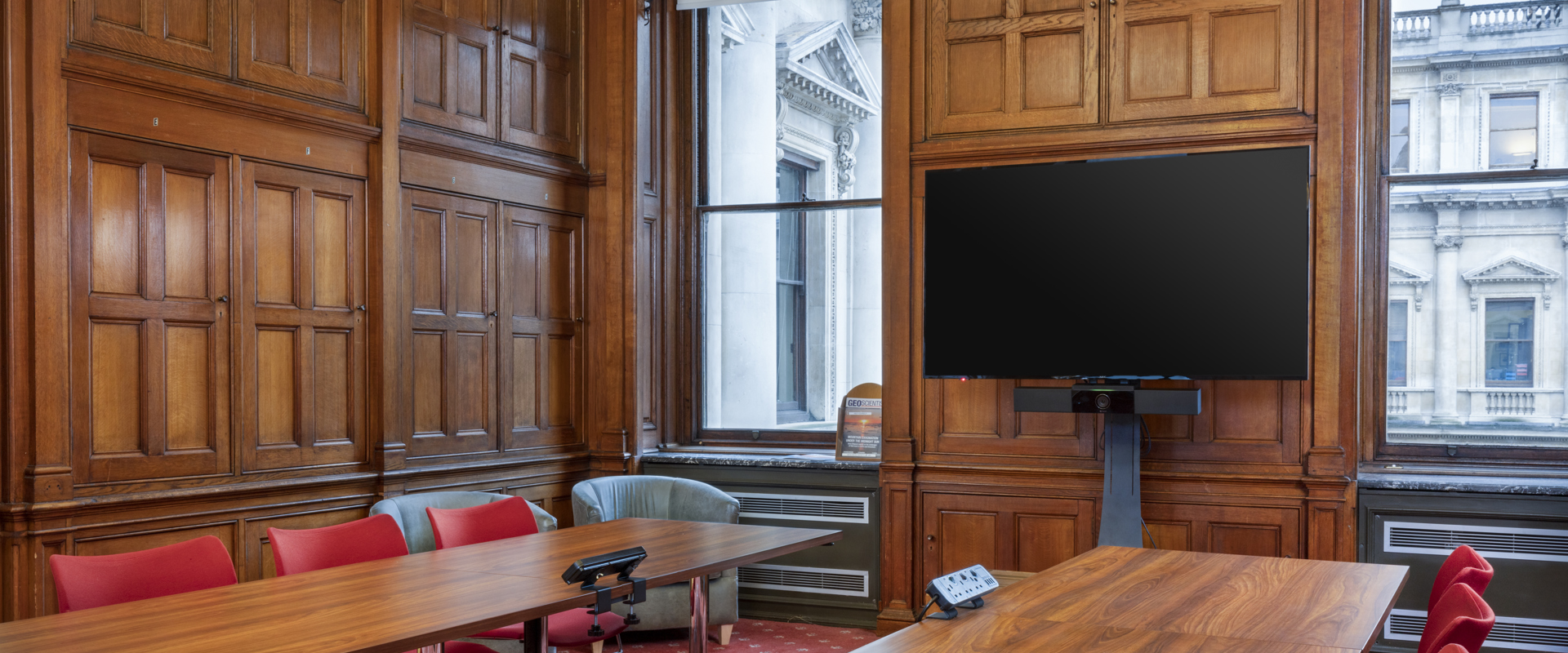 A spacious conference room with wooden paneling, a large oval table, red chairs, and a wall-mounted screen.