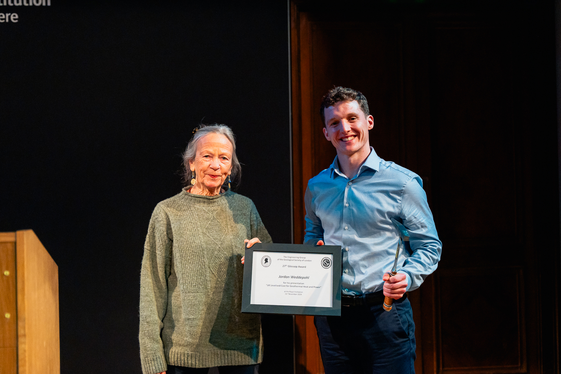 Two people stand on a stage. The older woman wears a grey sweater and smiles softly. The younger man holds a certificate and a small object, smiling proudly.