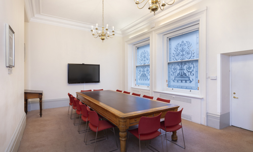 A spacious meeting room featuring a long wooden table, red chairs, decorative windows, in a bright setting.