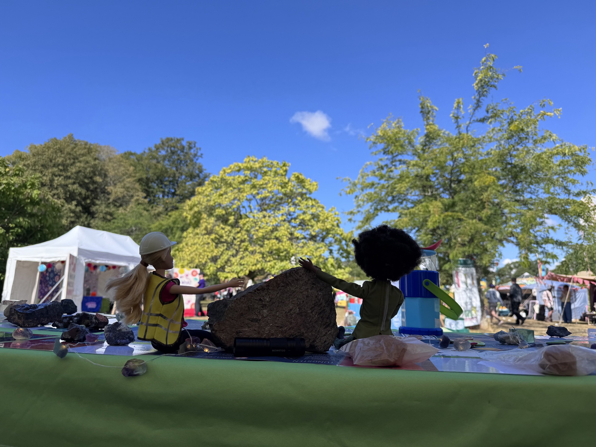 Two geologist barbies sit on a table with rocks and minerals, with a backdrop of the blue sky
