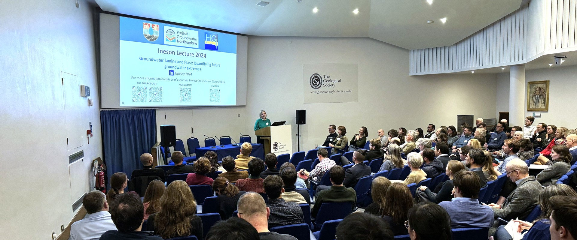Audience attending a lecture in a spacious venue, with a presenter at a podium and a large screen displaying the event's title and details.