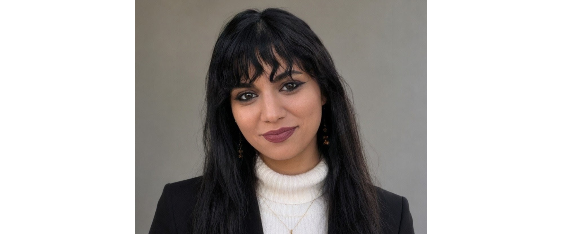 Close-up portrait of a person with long dark hair and bangs, wearing a black blazer over a white high-neck sweater and a small gold pendant, against a plain light background.