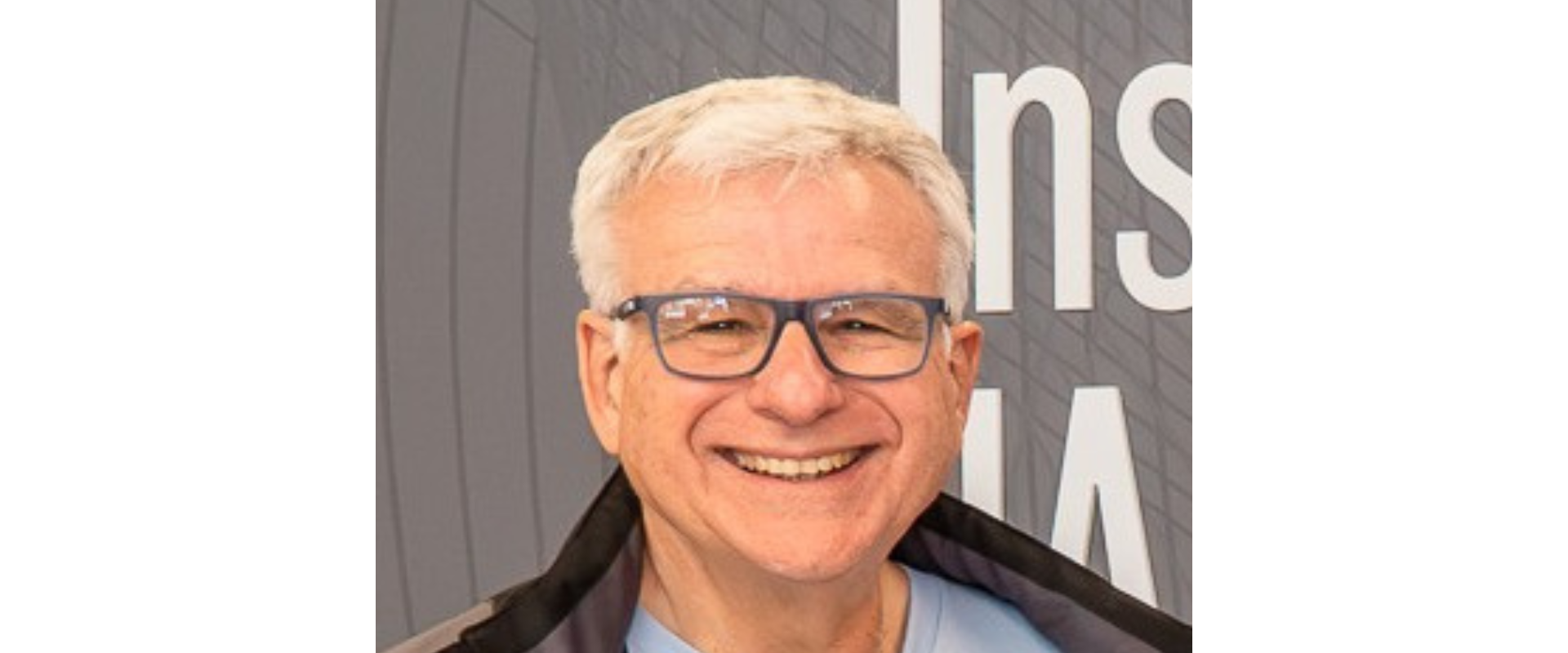 Head-and-shoulders portrait of a person with short light-colored hair and dark-framed glasses standing in front of a gray exhibition-style backdrop. The person is wearing a light blue shirt and a dark jacket draped around the shoulders, and is holding a document or brochure. Large partial white letters are visible on the background behind the person. The image is evenly lit and cropped closely around the upper body.