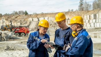 Three workers in helmets and uniforms discuss plans on a tablet at a quarry site, with machinery and stone piles in the background.