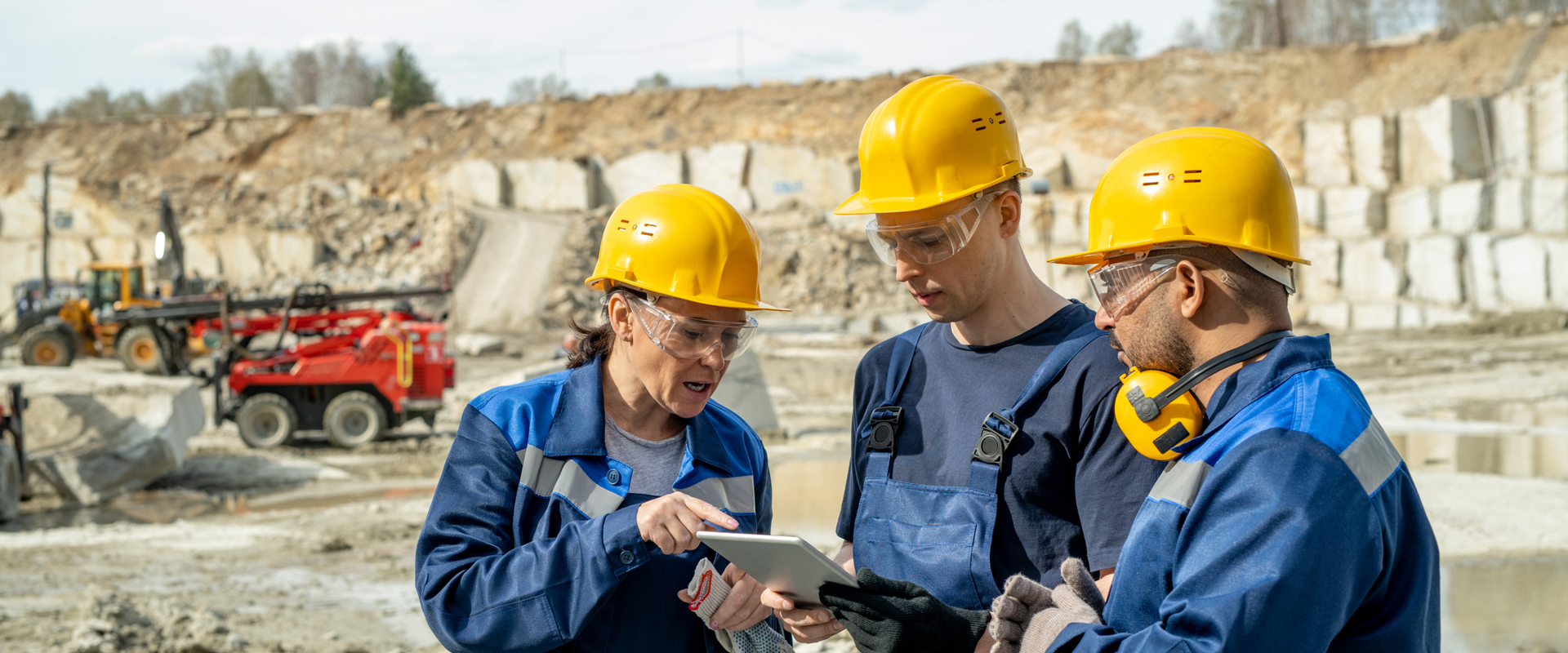 Three workers in helmets and uniforms discuss plans on a tablet at a quarry site, with machinery and stone piles in the background.