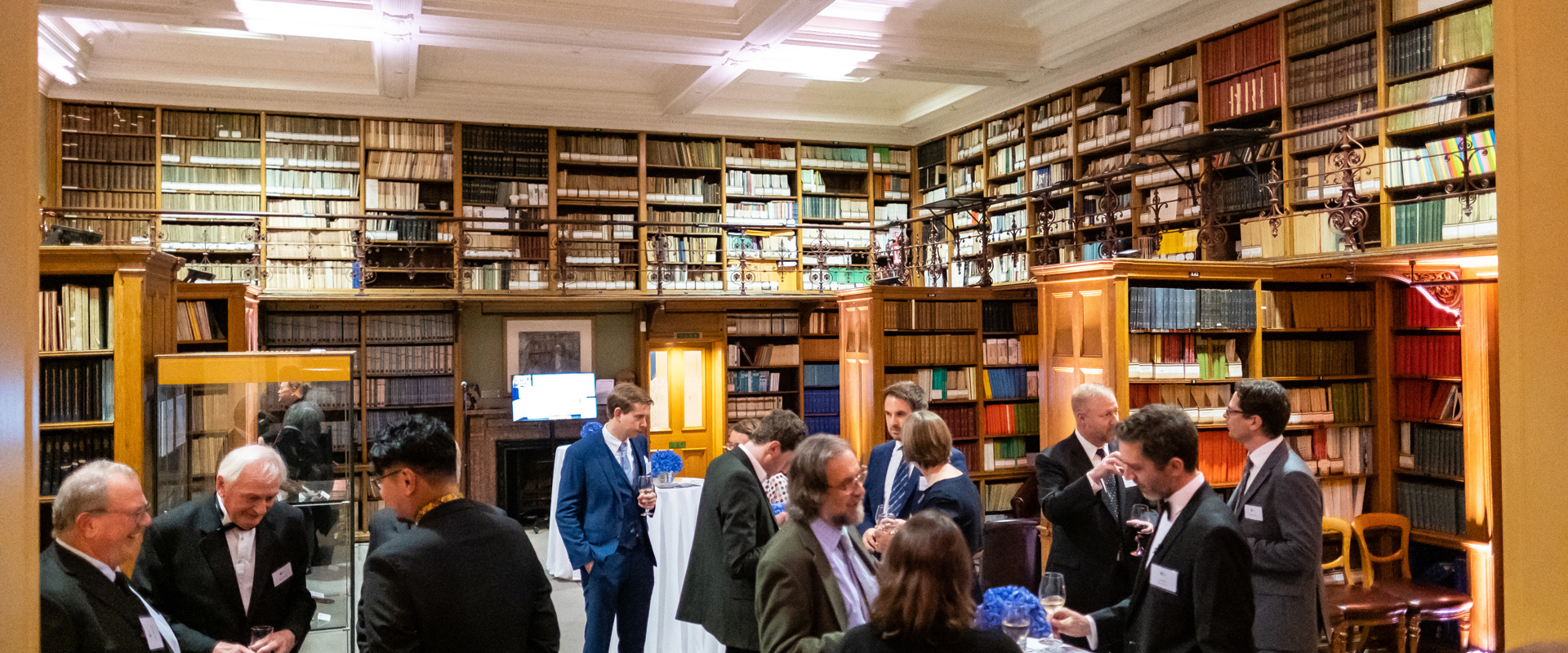 people stood by white linen covered tall tables holding drinks within a period library setting