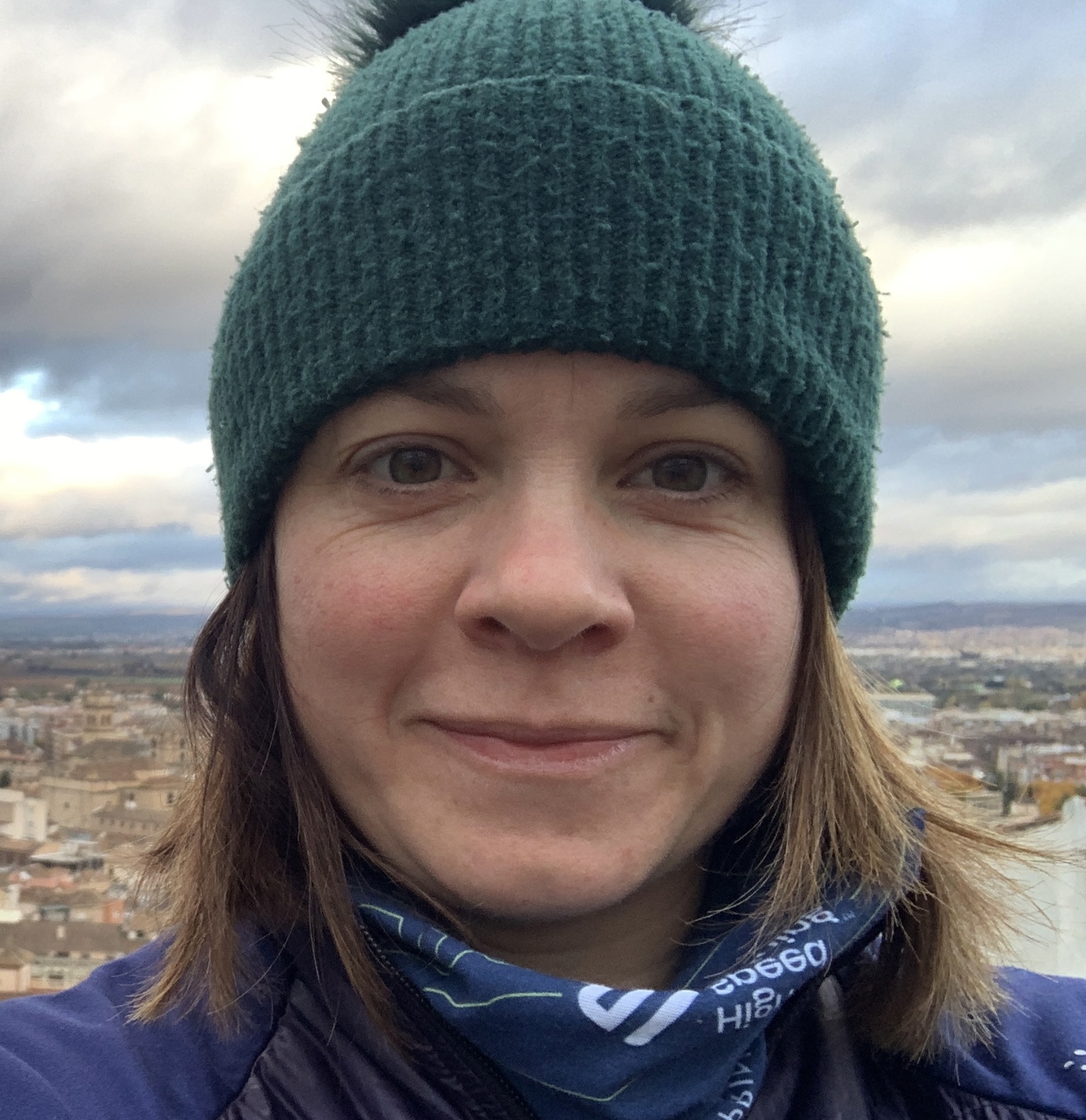 A person wearing a green knit hat and blue neck gaiter is outdoors with a cityscape and cloudy sky in the background.