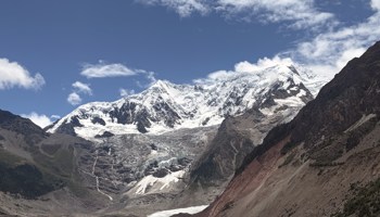 Snow-capped mountain peaks rise above a glacier-fed valley, with rocky slopes on either side and a calm lake containing small icebergs in the foreground. White clouds drift across a deep blue sky, and the glacier extends down from the mountains toward the water.