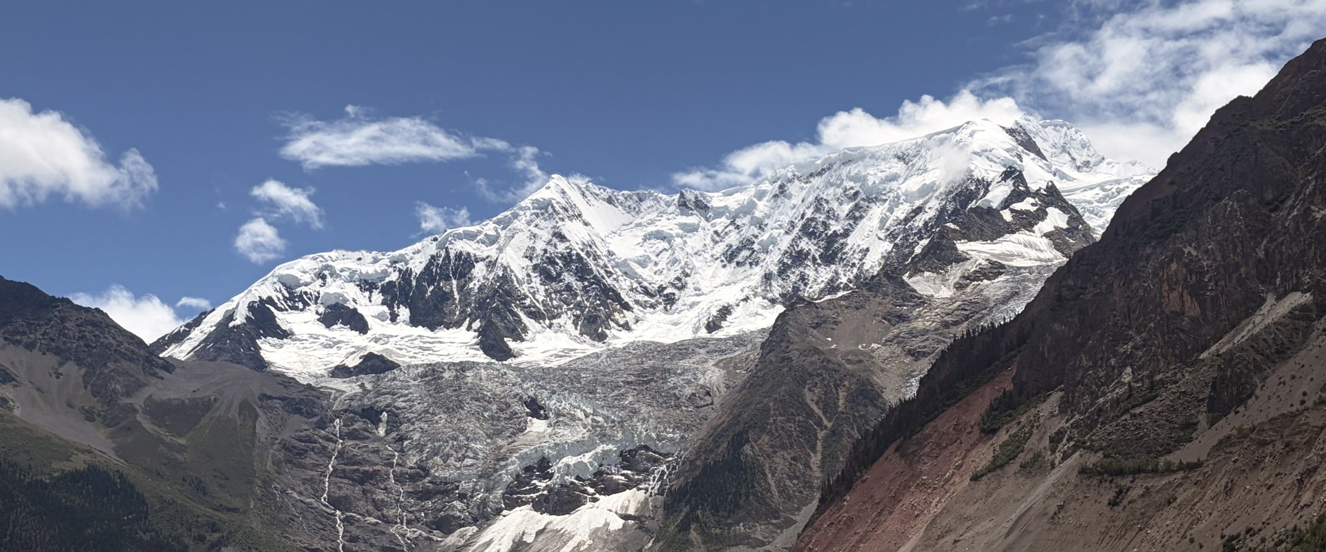 Snow-capped mountain peaks rise above a glacier-fed valley, with rocky slopes on either side and a calm lake containing small icebergs in the foreground. White clouds drift across a deep blue sky, and the glacier extends down from the mountains toward the water.