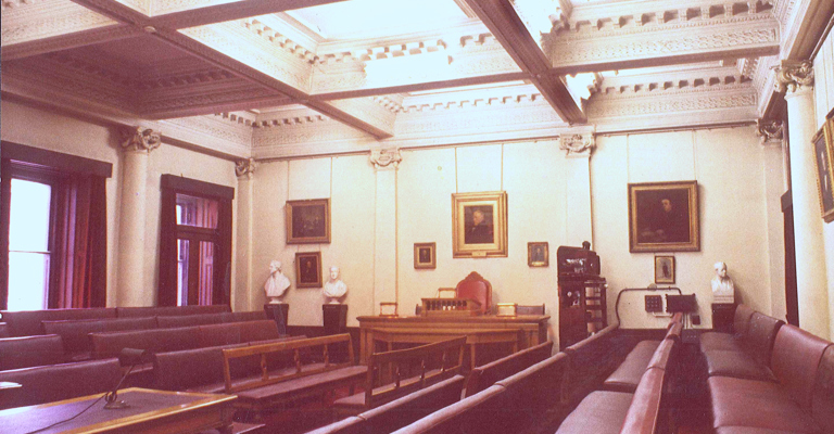 an old fashioned meeting room with dark burgundy sofas either side, tall sash windows and portraits on the walls