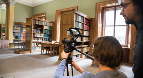 A person and an assistant recording a video in the Lower Library, surrounded by books and shelves, focused on their camera.