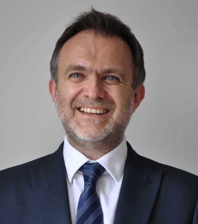 White man with grey hair and grey beard wearing a white shirt with tie and a navy suit jacket