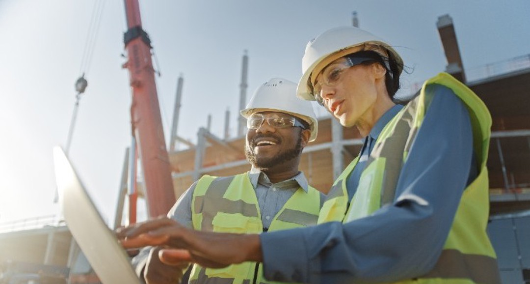 Two people in hardhats on site looking at a laptop