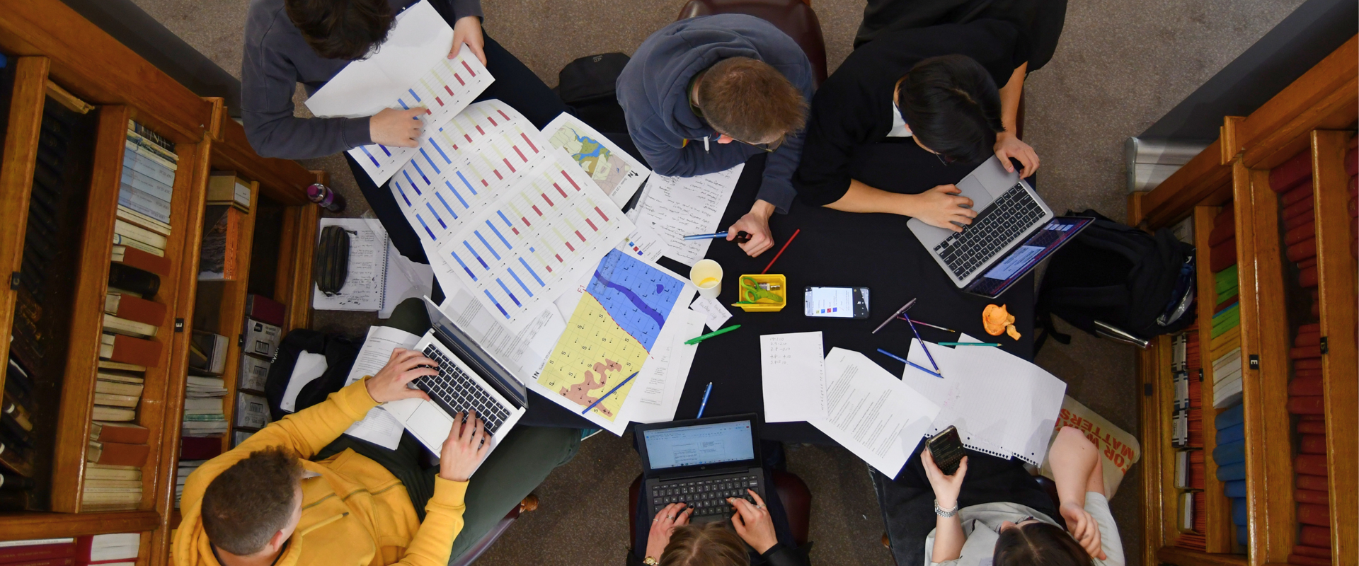 Bird eye shot of five kids working on a library desk with papers, maps and computers