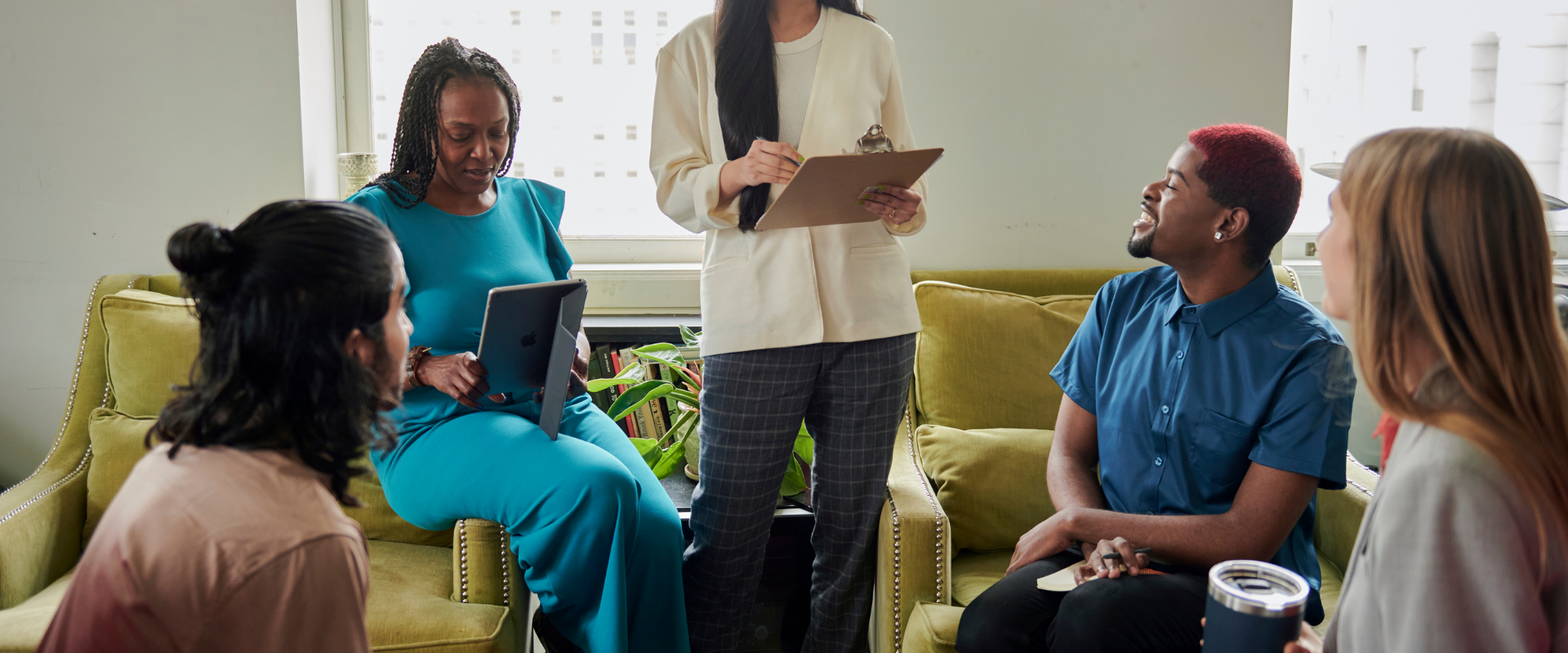 A woman stands holding a clipboard, leading a discussion with four colleagues seated on armchairs in a bright office.