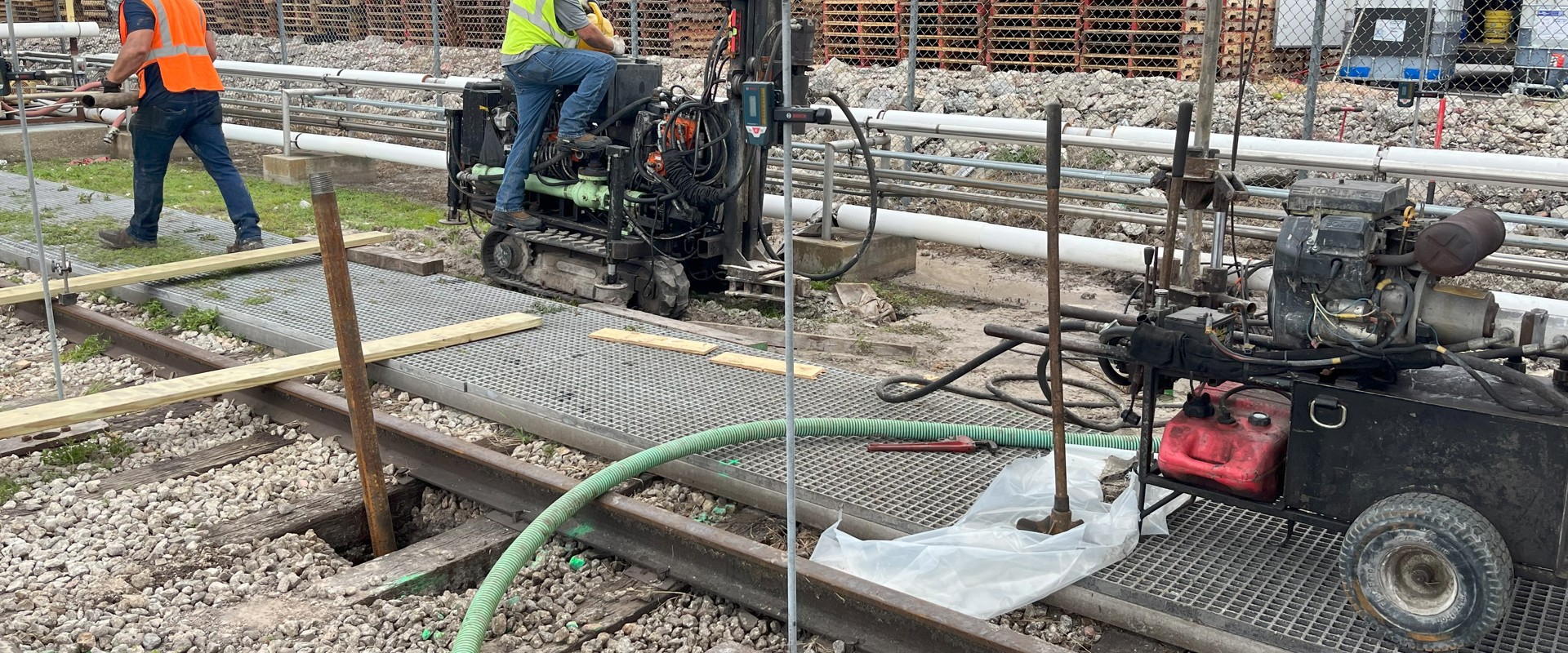 Two construction workers wearing safety gear operate drilling equipment along a set of railway tracks. One worker is seated on a small drilling rig, while another stands nearby. The scene includes hoses, machinery, and stacks of wooden pallets in the background.