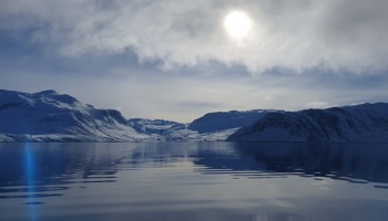 Snow-covered mountains rise from the shoreline of a calm, dark-blue fjord beneath a cloudy Arctic sky. The sun glows through a break in the clouds, casting a soft reflection across the still water, while distant snowy valleys and rugged peaks stretch across the horizon. A faint blue lens flare appears near the left edge of the image.