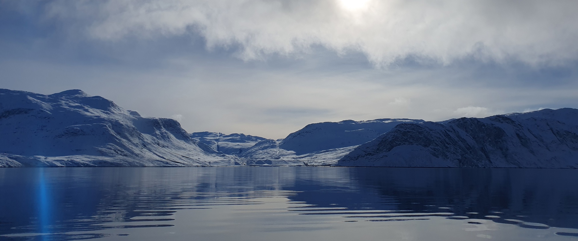 Snow-covered mountains rise from the shoreline of a calm, dark-blue fjord beneath a cloudy Arctic sky. The sun glows through a break in the clouds, casting a soft reflection across the still water, while distant snowy valleys and rugged peaks stretch across the horizon. A faint blue lens flare appears near the left edge of the image.