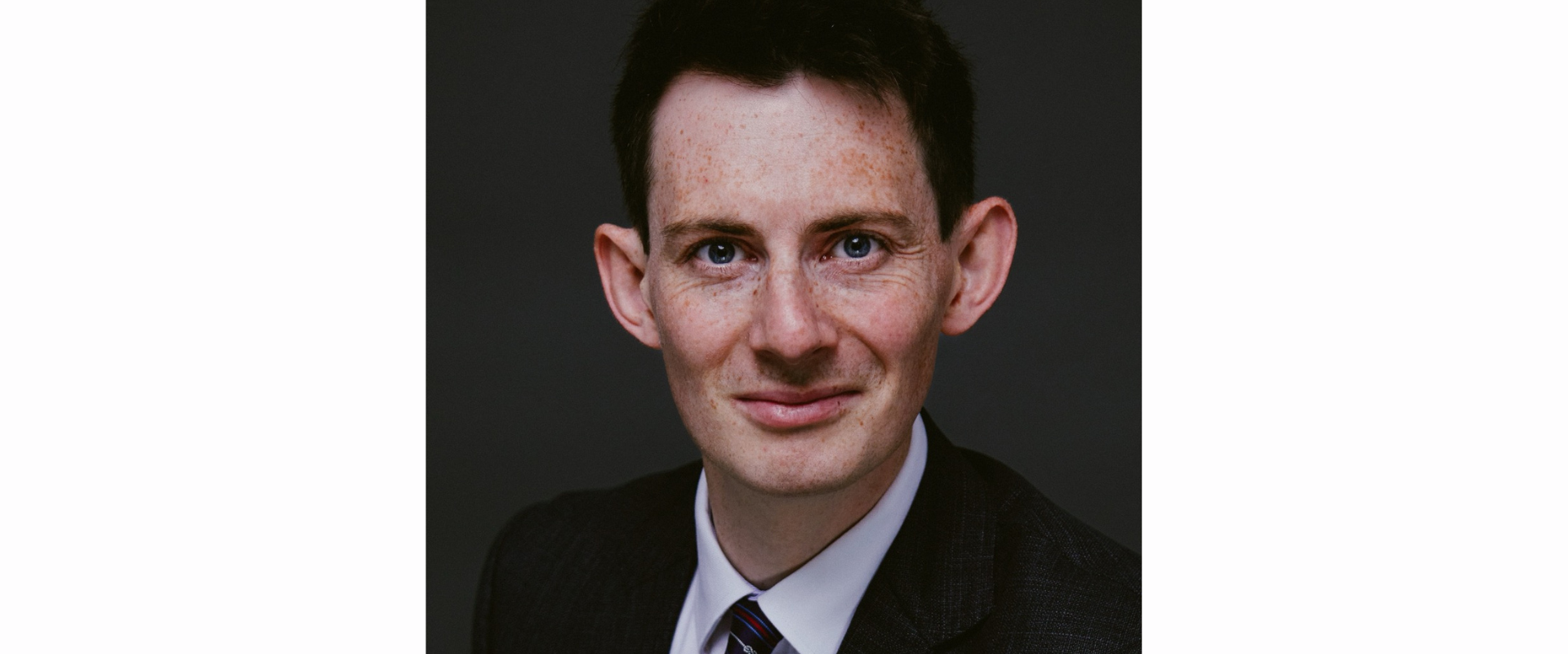 A person wearing a dark textured suit jacket, a white collared shirt, and a striped tie, posed against a plain dark background.
