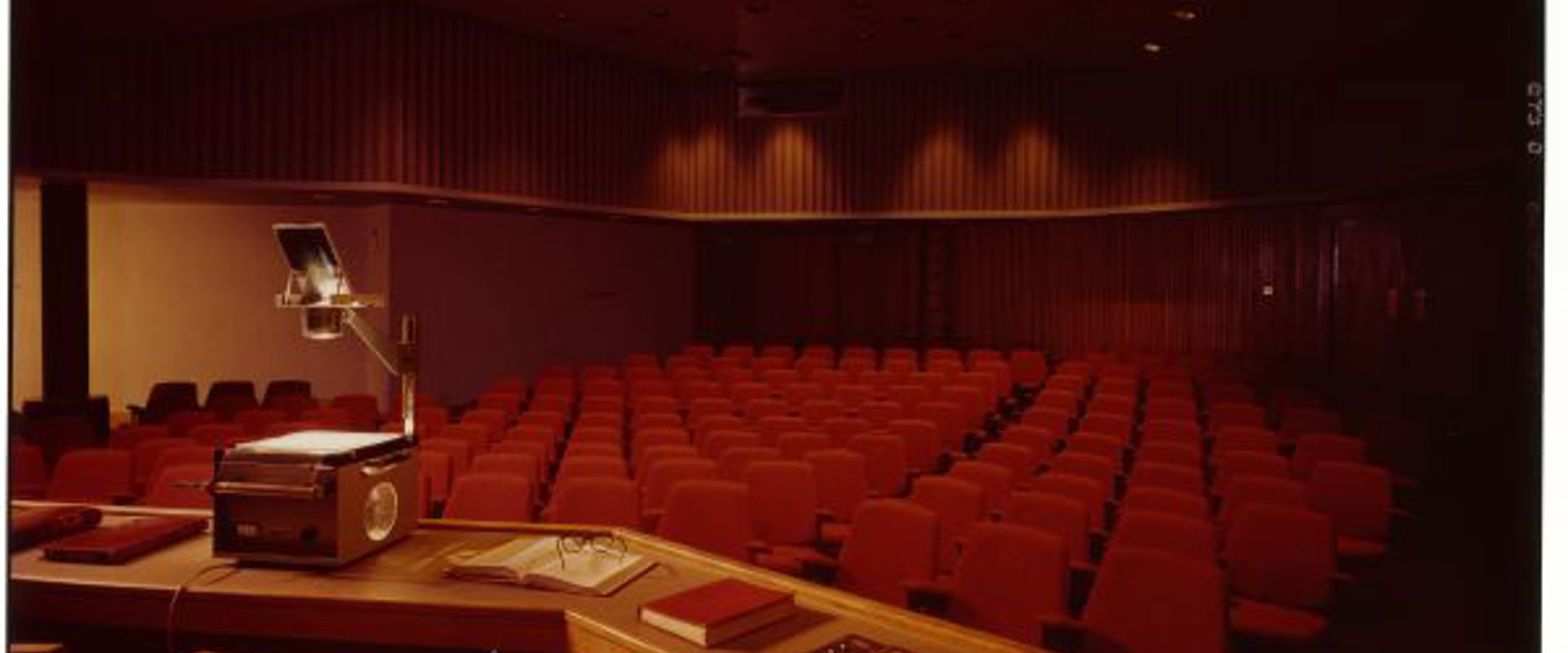 Lecture theatre with red seating and an overhead projector on a desk with laid out books and notebooks