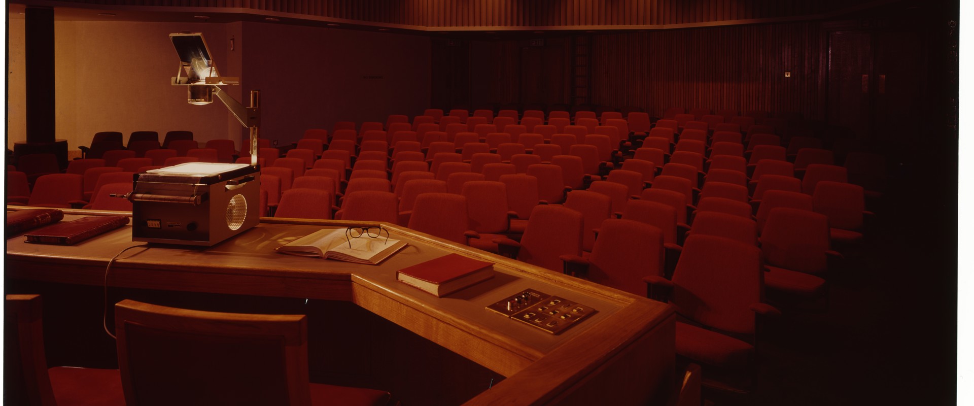 Lecture theatre with red seating and an overhead projector on a desk with laid out books and notebooks