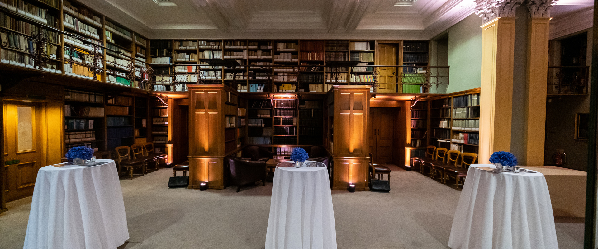 a wood panelled period library space with book shelves and three tall tables covered in white tablecloths, ready for an event