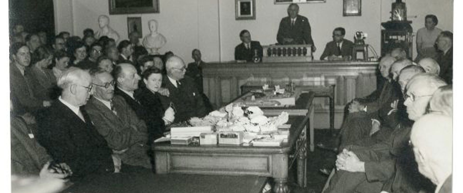 Old photograph of individuals seeting in a room facing each other durint a meeting