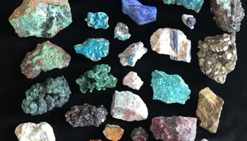 A flat lay of around thirty rough, unpolished mineral specimens arranged neatly on a black background. The rocks vary in size and shape and display a wide range of colours, including turquoise, teal, deep blue, pale green, white, pink, purple, yellow, orange, and metallic gold. Some pieces appear crystalline or granular, while others are more solid and blocky, highlighting the varied textures and natural forms of the minerals.