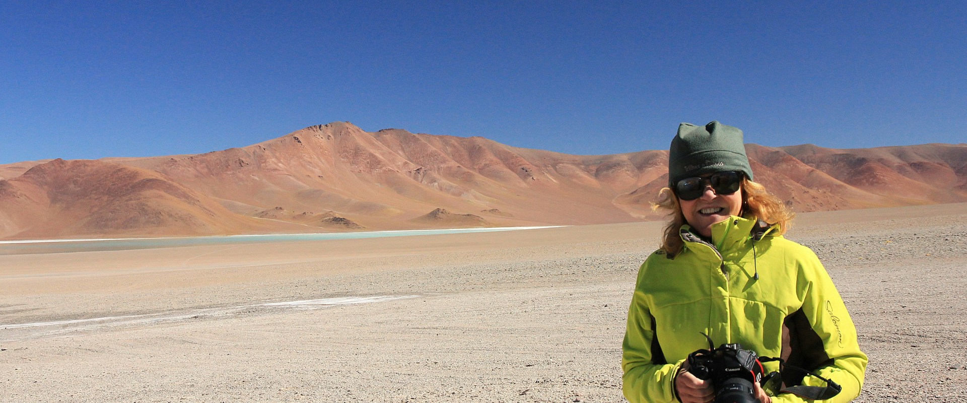 woman with high vis coat and green beanie hat stood in front of a desert are with mountains in the background