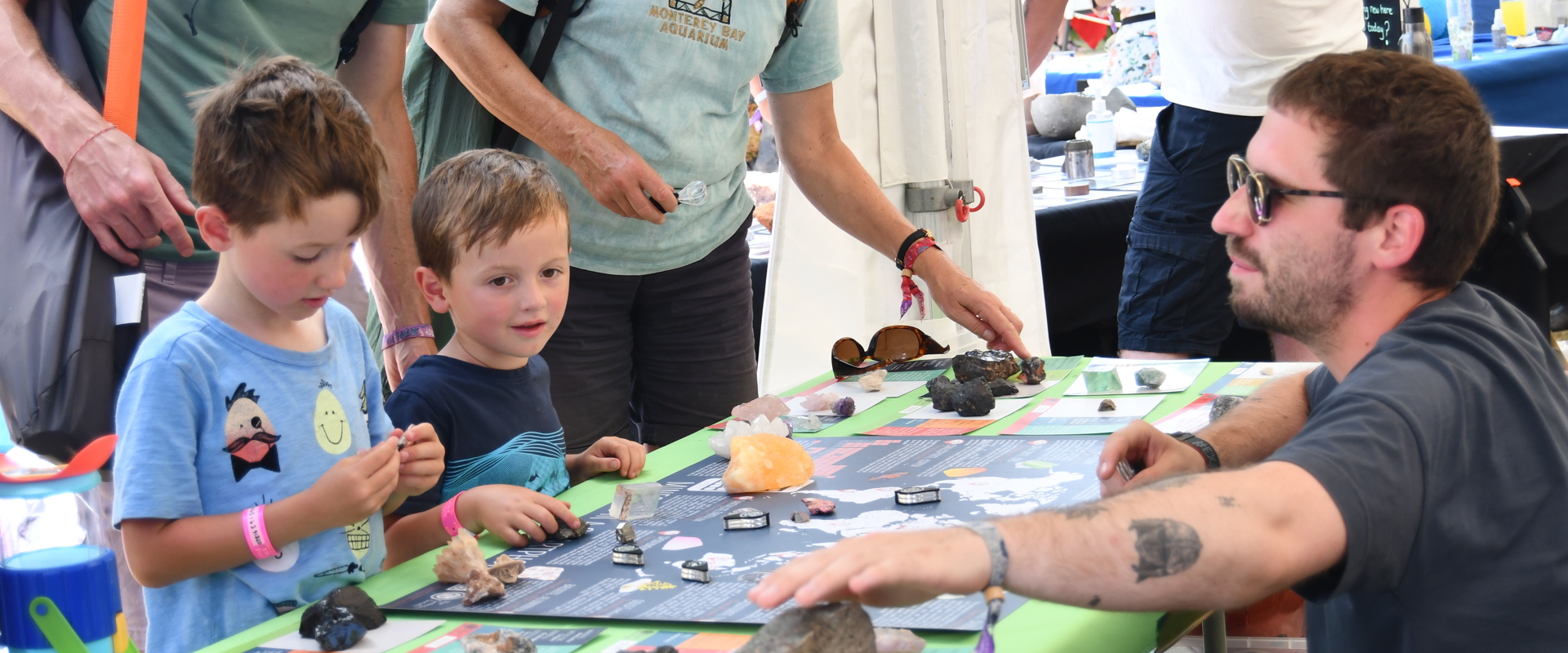 Two kids and two adults interact with a science communicator and rocks on a table during a festival