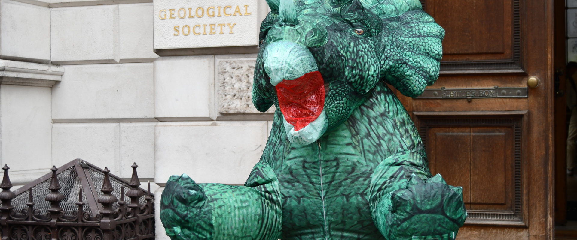 someone in an inflatable dinosaur suit stood in front of a building with a plaque reading the geological society