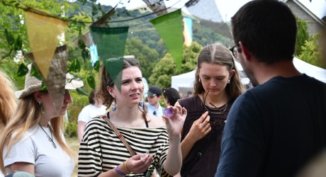 Three women and one man stand and talk outdoors near colourful pennant banners, with trees and tents visible in the background.