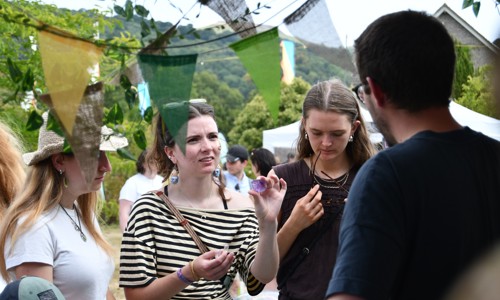 Three women and one man stand and talk outdoors near colourful pennant banners, with trees and tents visible in the background.