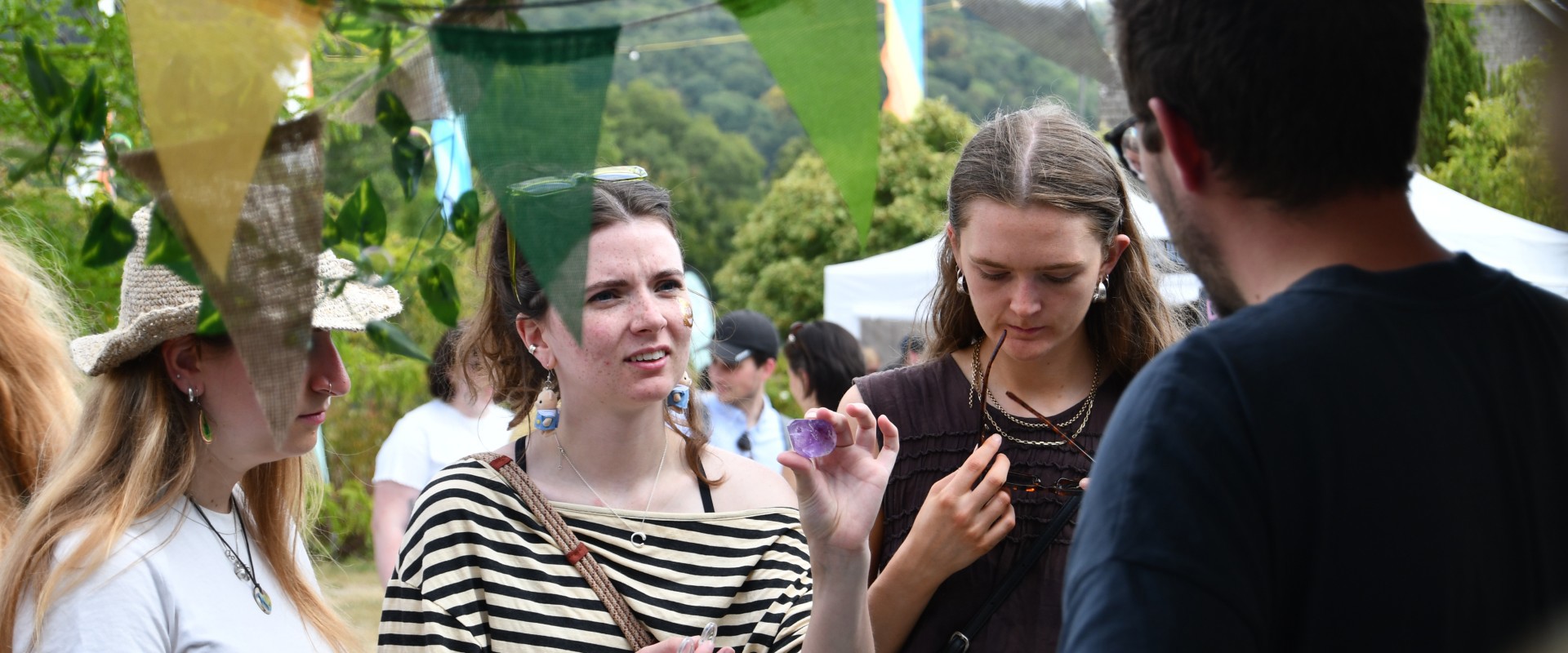 Three women and one man stand and talk outdoors near colourful pennant banners, with trees and tents visible in the background.