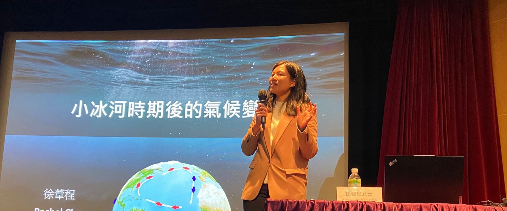 an Asian woman speaking into a microphone stood at a curtain covered table with a projector screen behind her