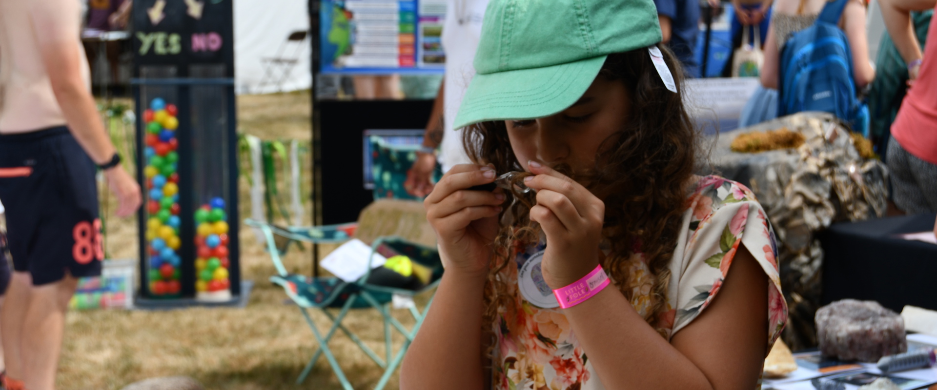 Girl holding a magnifying glass in a festival environment, surrounded by other people and in front of a table with rocks
