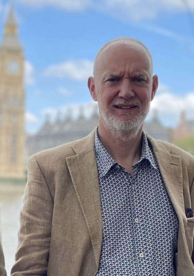 bald white man with grey beard wearing a beige jacket and pattened shirt