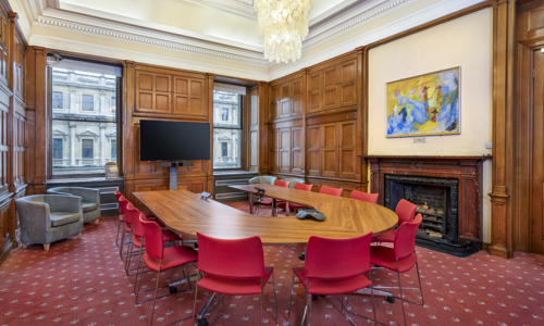 A spacious conference room with wooden paneling, a large oval table, red chairs, and a wall-mounted screen.