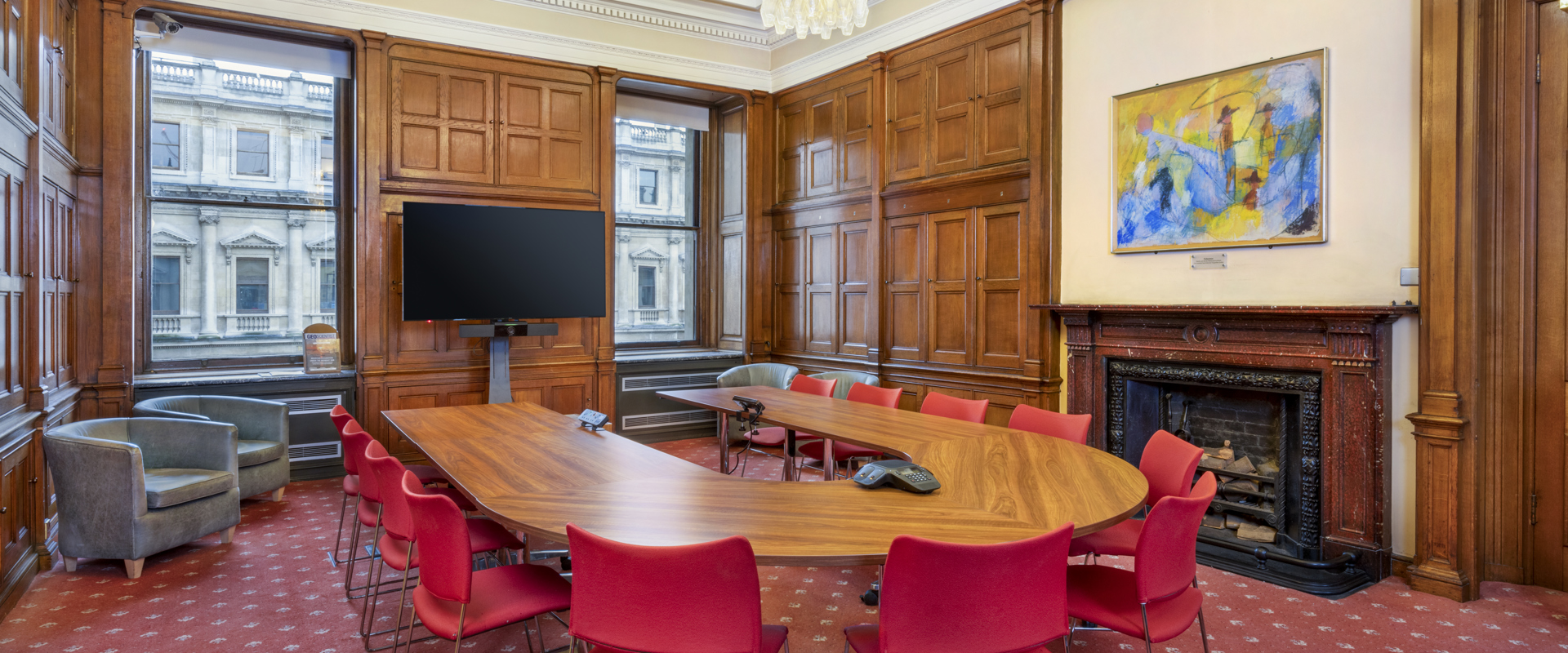 A spacious conference room with wooden paneling, a large oval table, red chairs, and a wall-mounted screen.