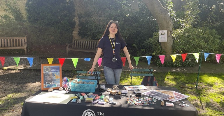 young white woman with long brown hair smiling whilst stood behind a table covered with a blue tablecloth with The Geological Society written on it