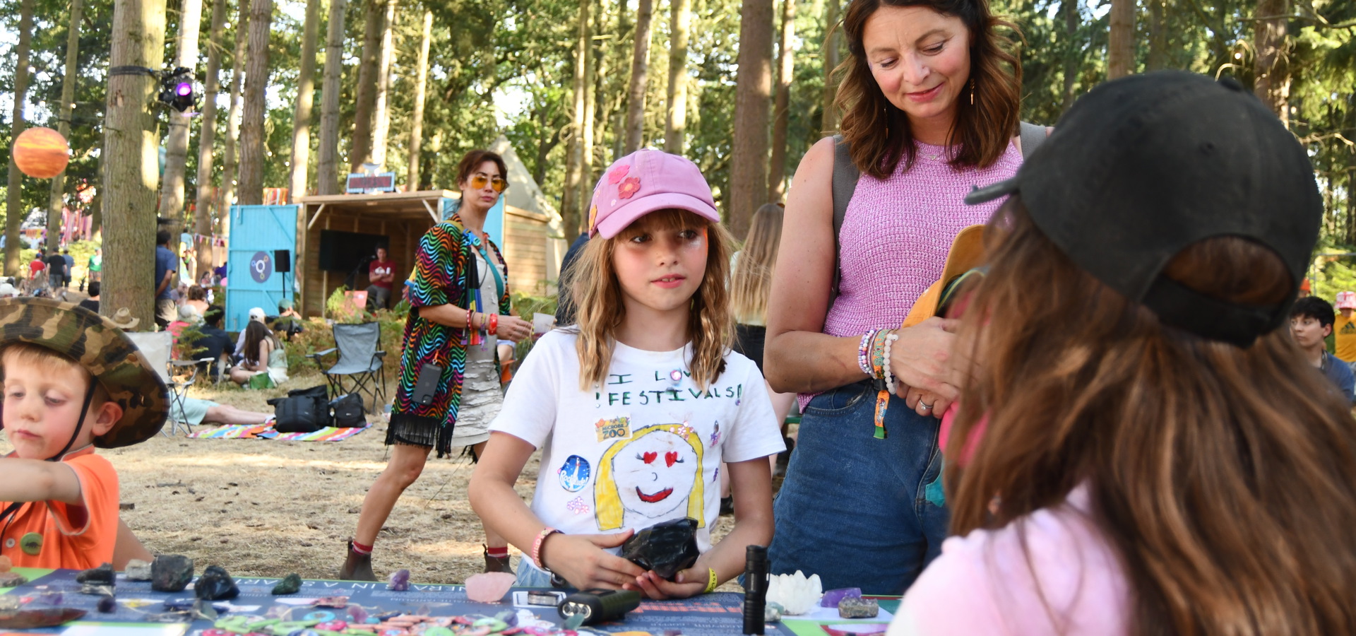 a small girl wearing a pink cap and white t shirt standing with a dark haired adult woman is listening to a woman sat at a table in a forest festival setting
