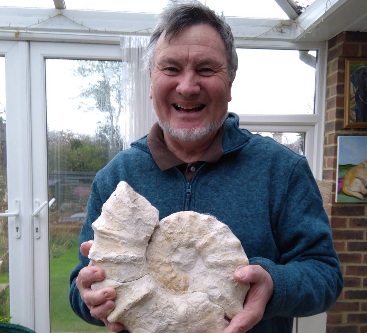 White man with grey hair and grey beard smiling holding a large ammonite fossil, stood inside a house with doors and a garden behind him