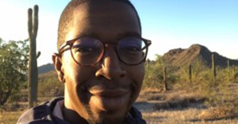 A man wearing glasses looking at the camera with a mountainous scene in the bakcground