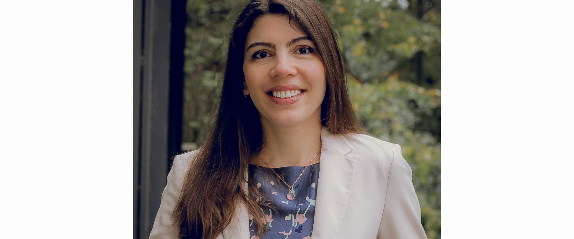 A person with long, straight brown hair is standing outdoors in front of green foliage. The person is wearing a light-coloured blazer over a patterned top with floral designs. The background is softly focused, showing trees and natural greenery.