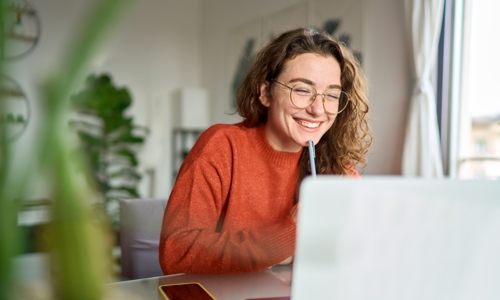 Young woman in glasses smiling at laptop with pen
