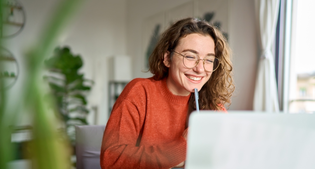 Young woman in glasses smiling at laptop with pen