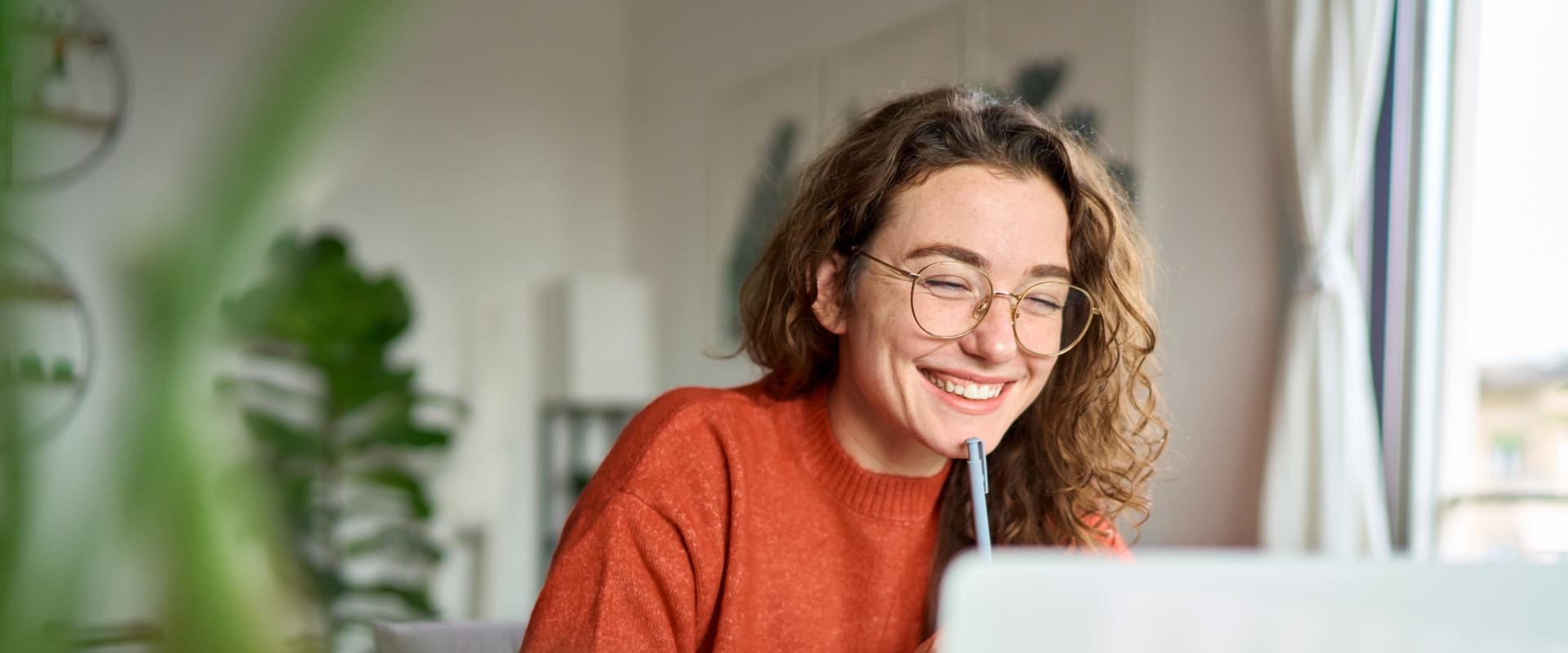 Young woman in glasses smiling at laptop with pen
