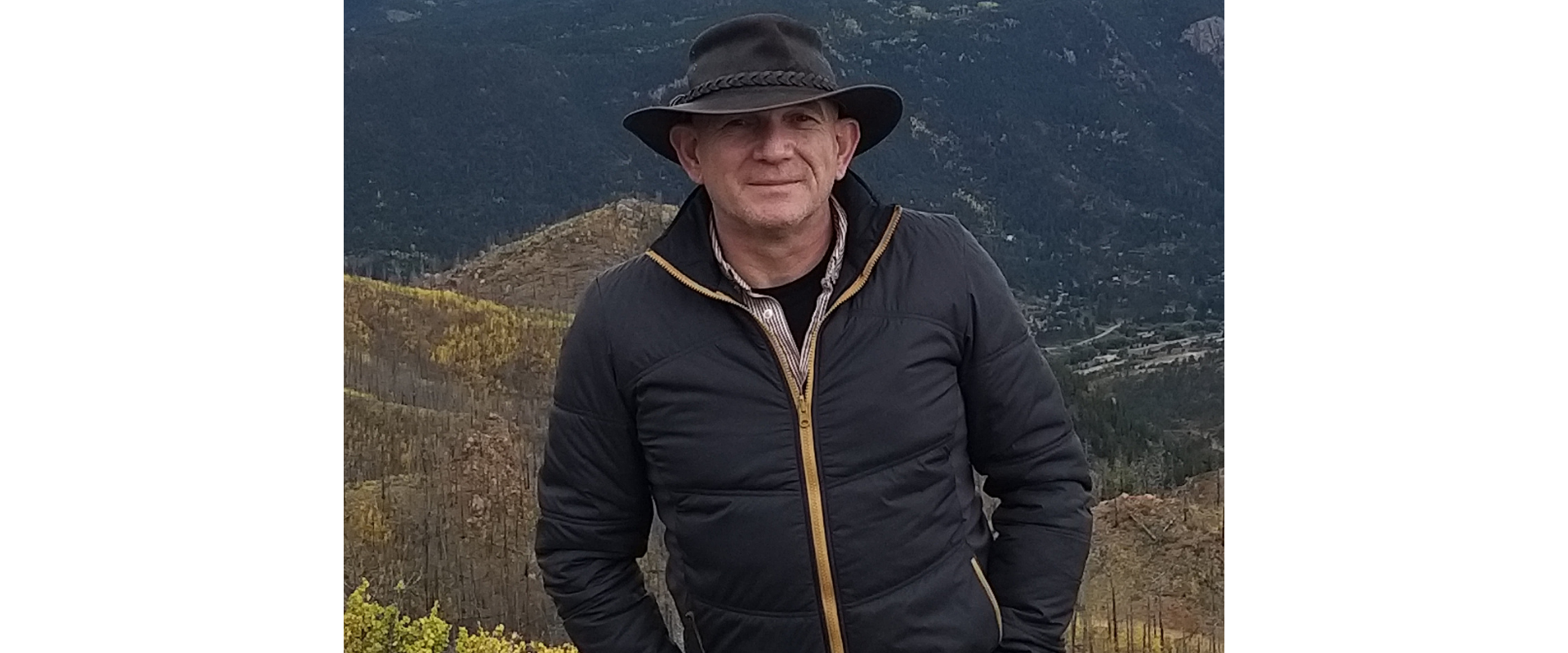 Person wearing a dark wide-brimmed hat and a dark insulated jacket standing on a mountain overlook, with forested hills and a valley stretching into the background. Autumn-colored shrubs and trees appear in the foreground under overcast light.